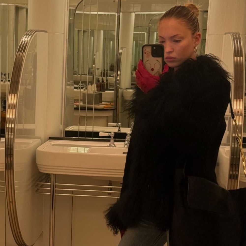 Lila Moss takes a mirror selfie in a bathroom wearing bootcut jeans with rounded-toe heels boots and a fur-trimmed jacket.