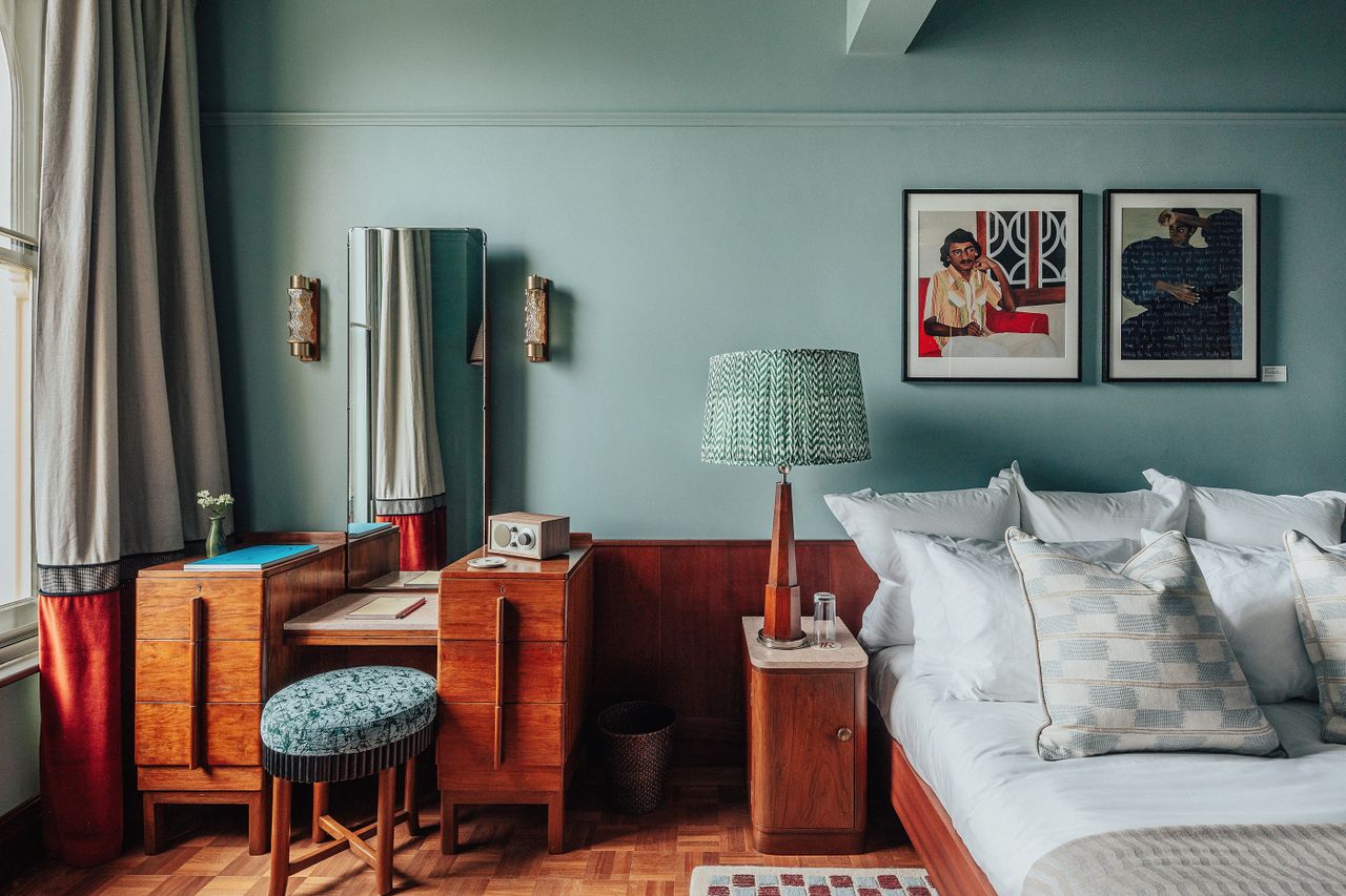 A layered, transitional hotel suite featuring a series of figurative, colorful artworks on its walls, patterned furniture, including rugs, armchairs, and sofas, split-color pillars, and a buttery, tile blue, and green palette.