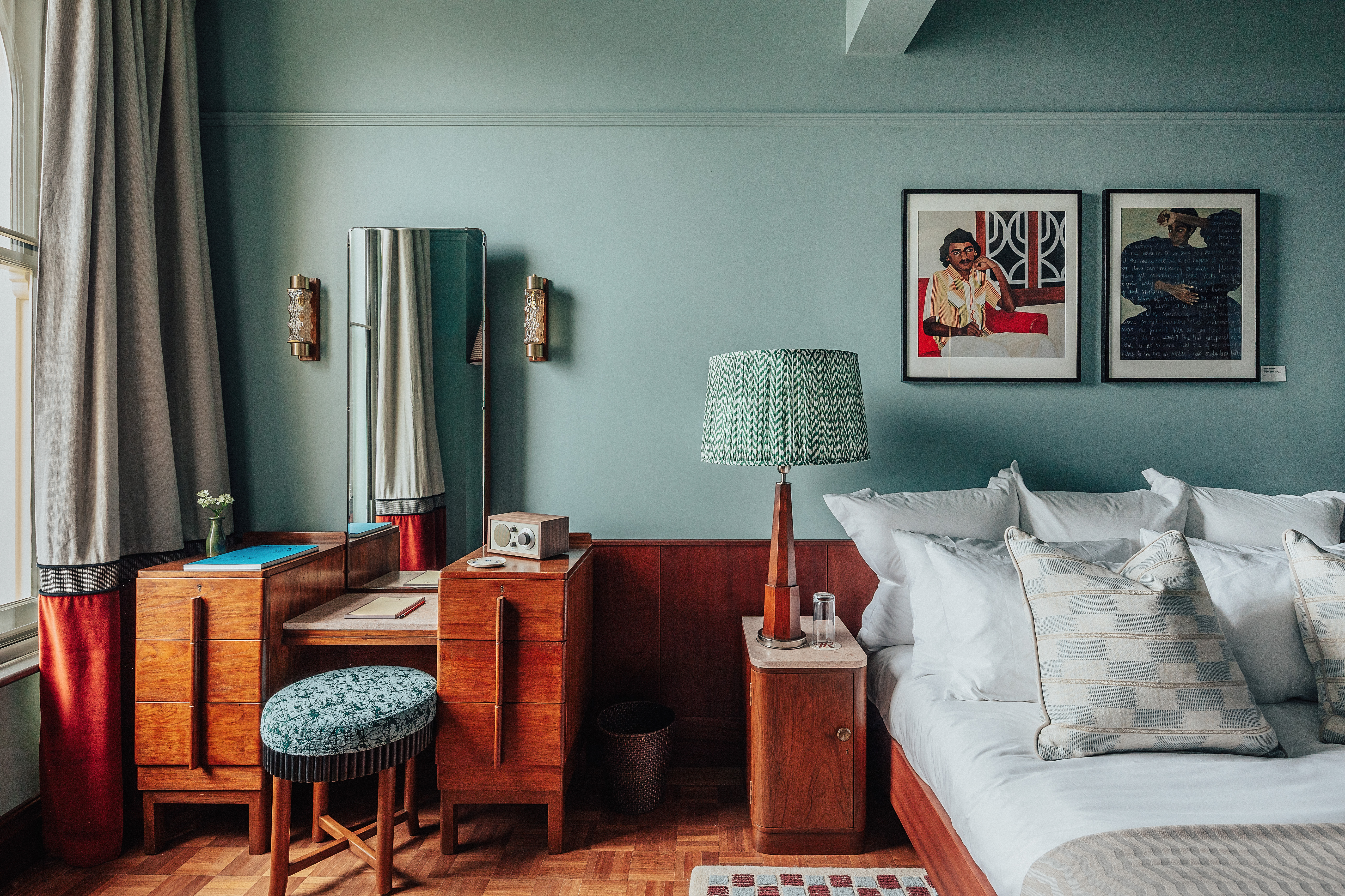 A layered, transitional hotel suite featuring a series of figurative, colorful artworks on its walls, patterned furniture, including rugs, armchairs, and sofas, split-color pillars, and a buttery, tile blue, and green palette.