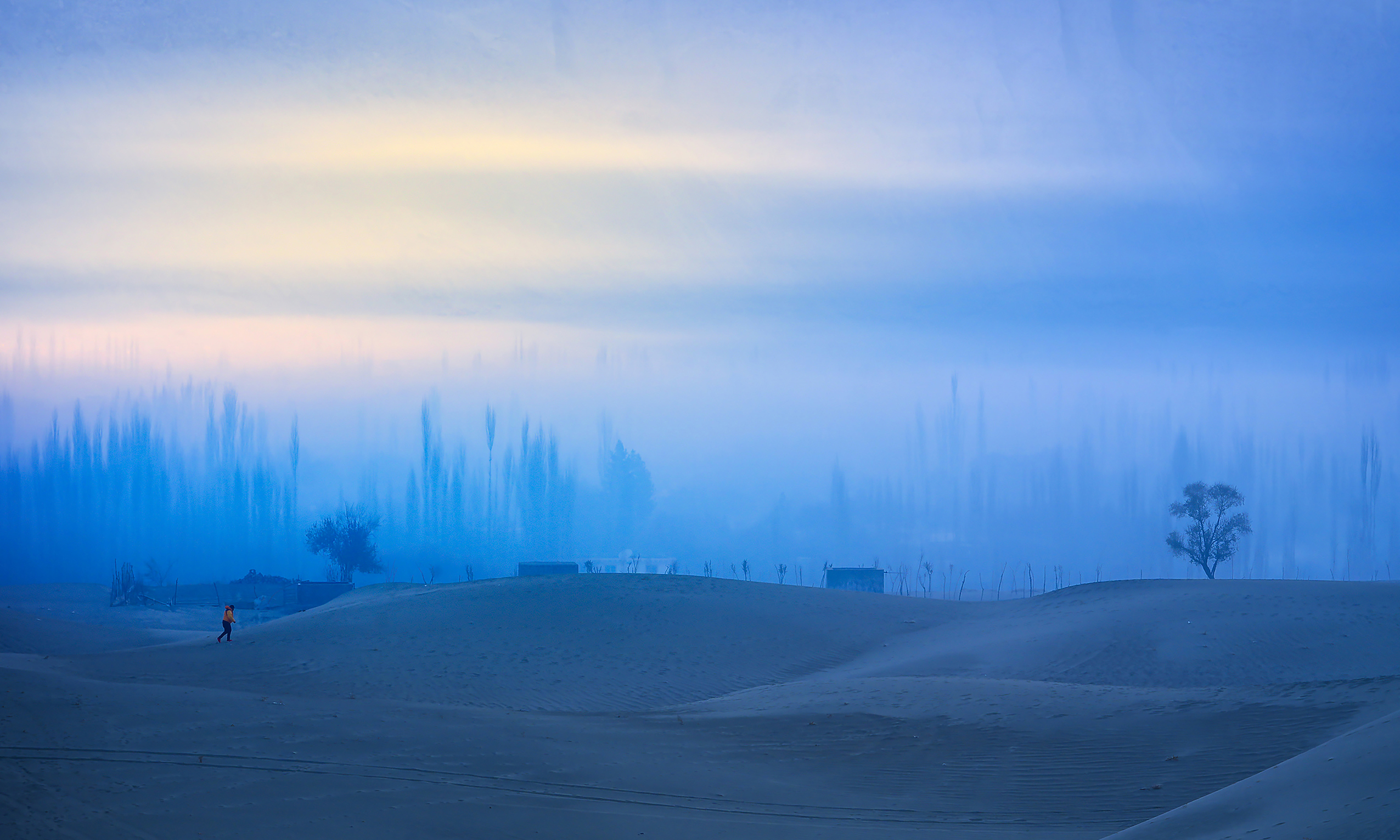 A lone figure walks on soft sand dunes under a blue, misty sky. Sparse trees and distant structures create a serene, dreamlike landscape at dawn