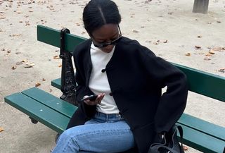  @sylviemus_ wearing a cream sweater, black cardigan, and jeans while sitting on a Paris park bench.