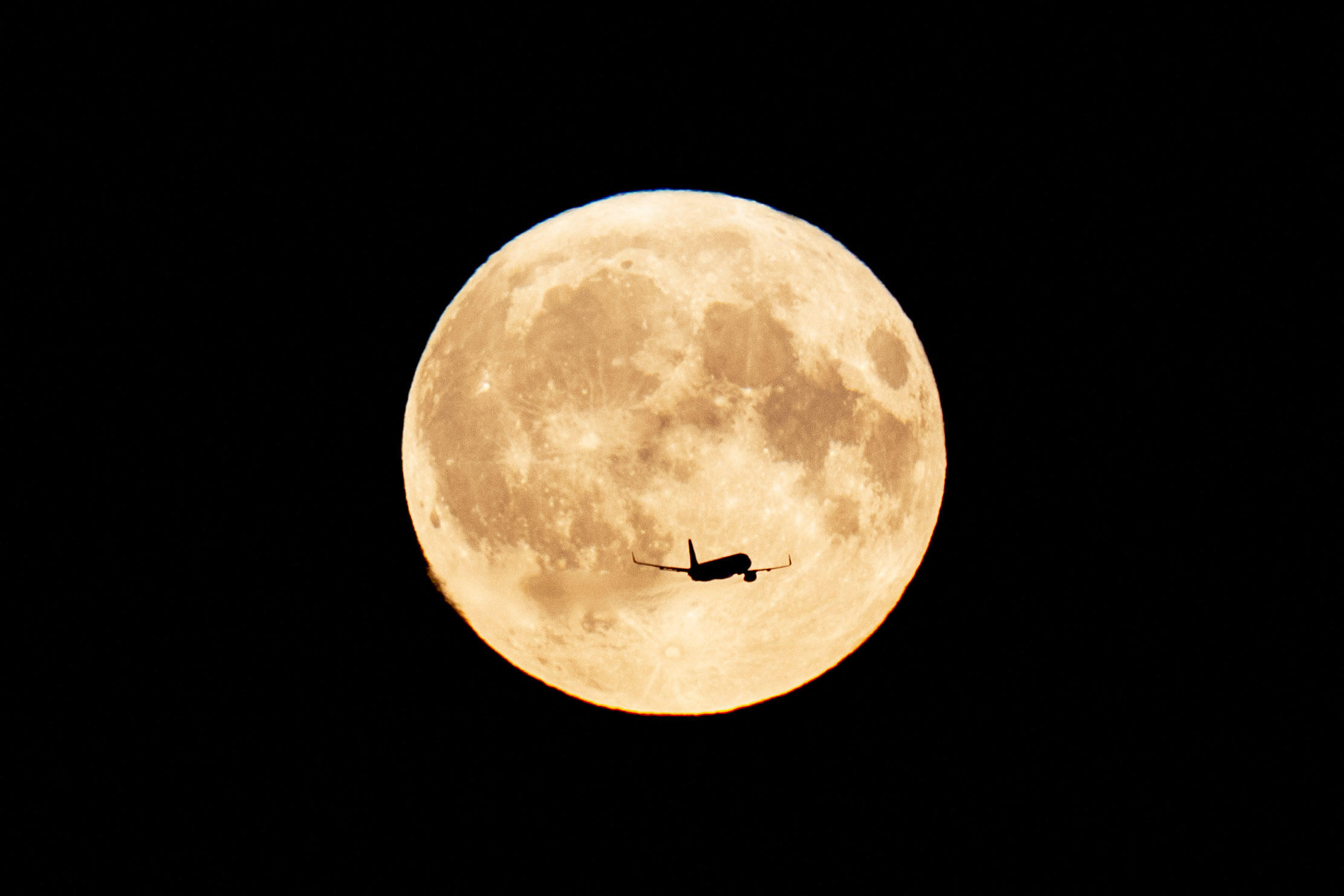 Bulan besar yang terang benderang di latar belakang dan di latar depan siluet pesawat terbang melintasi bulan.