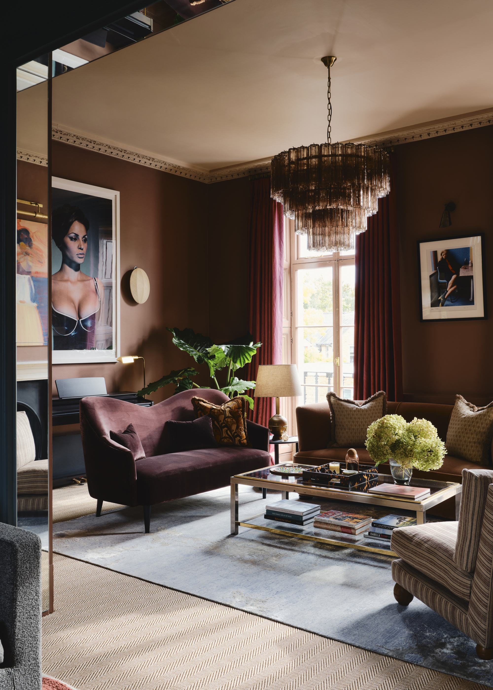 Image of a living room painted in a deep ochre yellow with crown molding on the ceilings. There is a purple velvet love seat and a dark ochre sofa in the room. On the floor is a light blue rug and a glass and gold coffee table.