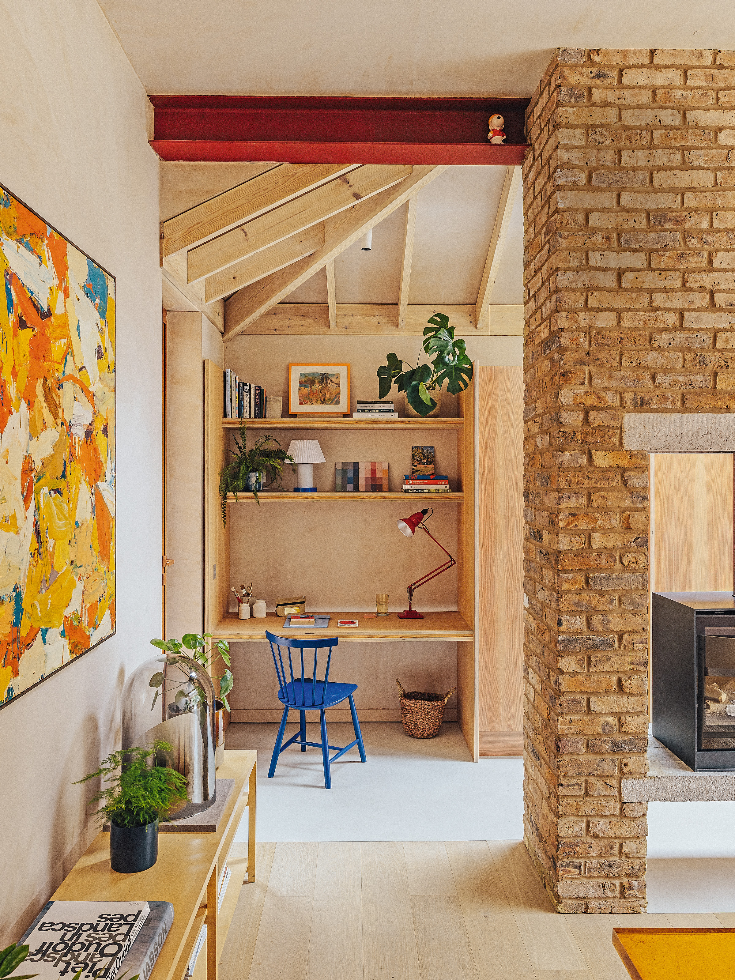 View past exposed brick fireplace to a built-in plywood desk and shelving area with a blue chair