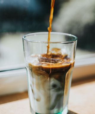 A glass coffee cup with white milk and ice in it, being topped up with brown coffee, stood on a brown window ledge with a blue window behind it