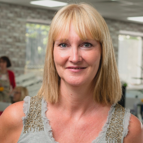 Image of a woman with short blonde hair and fringe. She's wearing a gray sleeveless top and the straps are beaded.