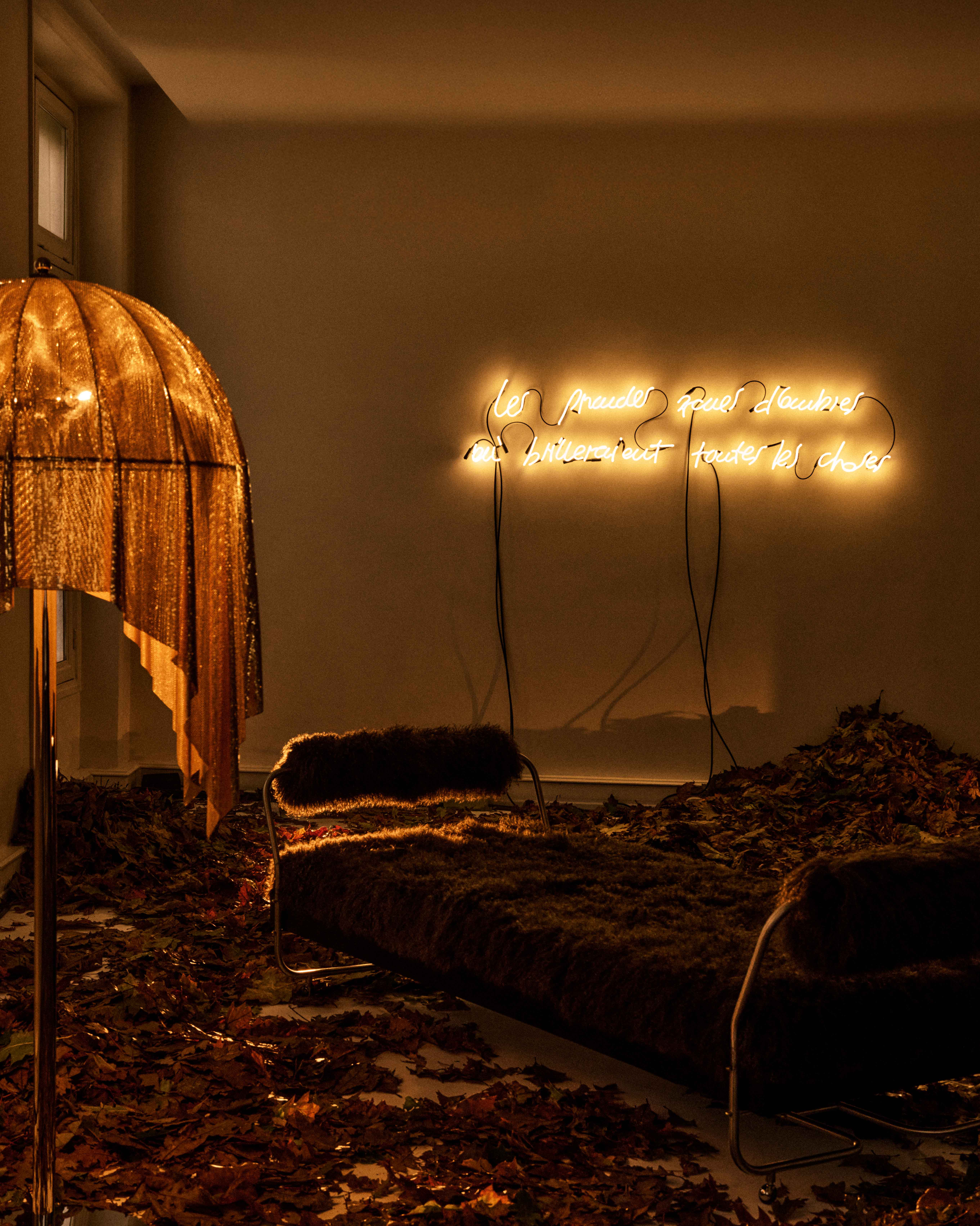 Dark room with petals on the floor, a lamp with an orange shade, a neon light with letters on the wall and a fur-upholstered daybed