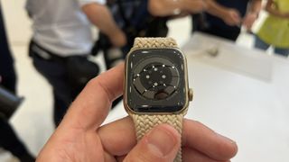 En hand som håller upp en Apple Watch 8 med ett beige textilarmband.