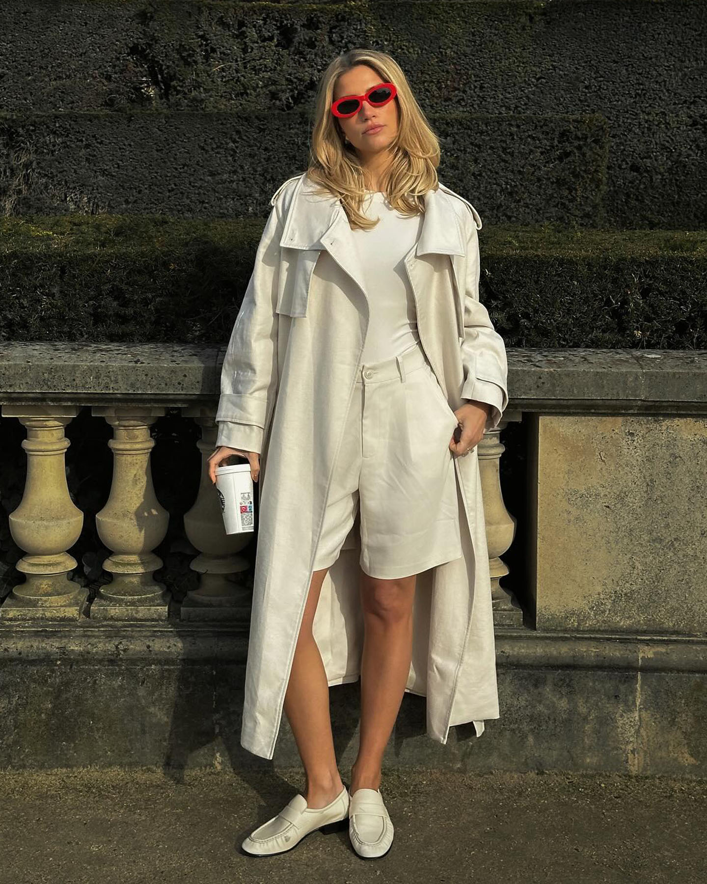 Influencer Cass Dimicco poses on a sidewalk in Paris wearing bright red oval sunglasses, a beige trench coat, white tee, beige Bermuda shorts, and beige flats from The Row.