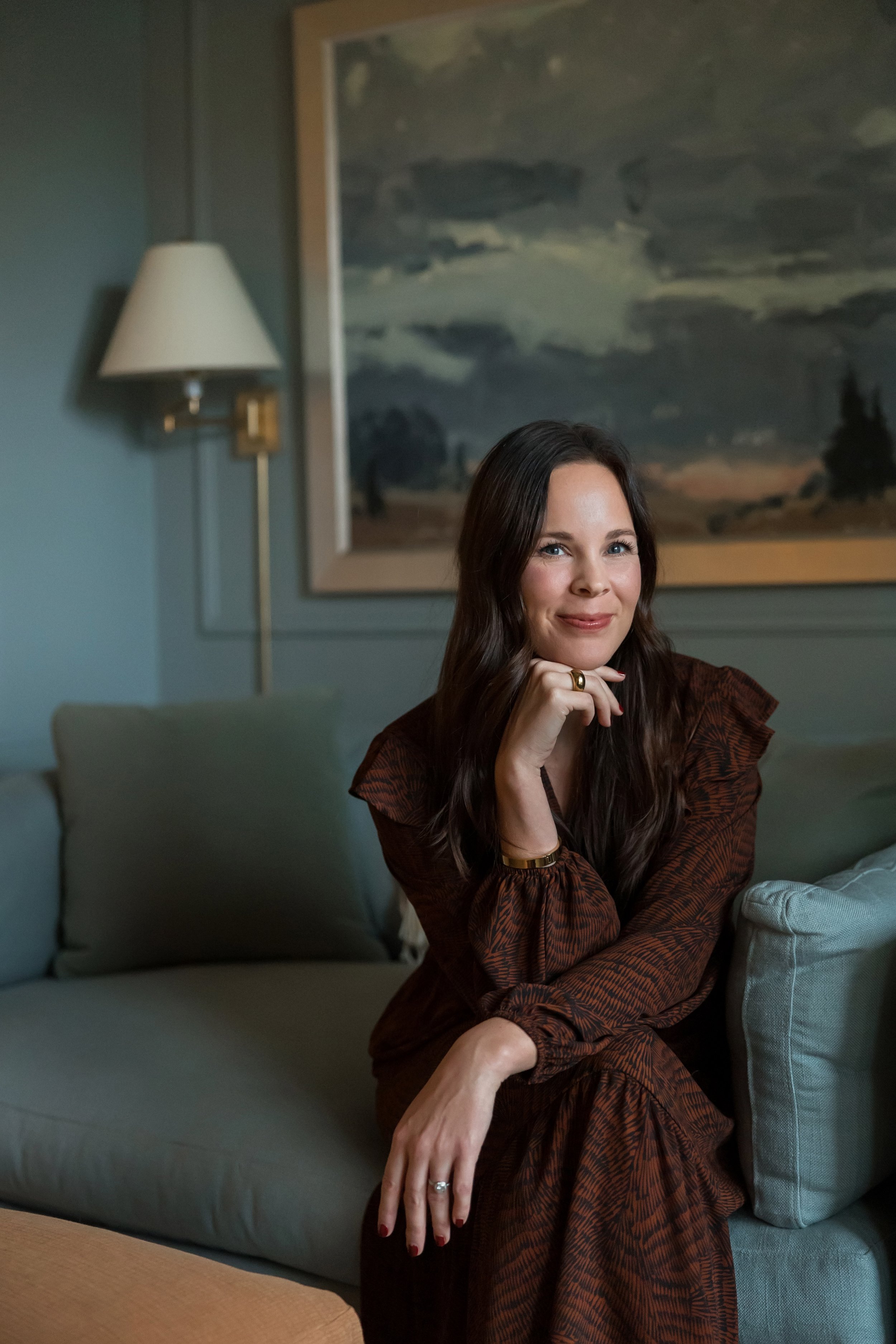 A brunette woman in a dress sits on a coach in a blue-gray colored room.