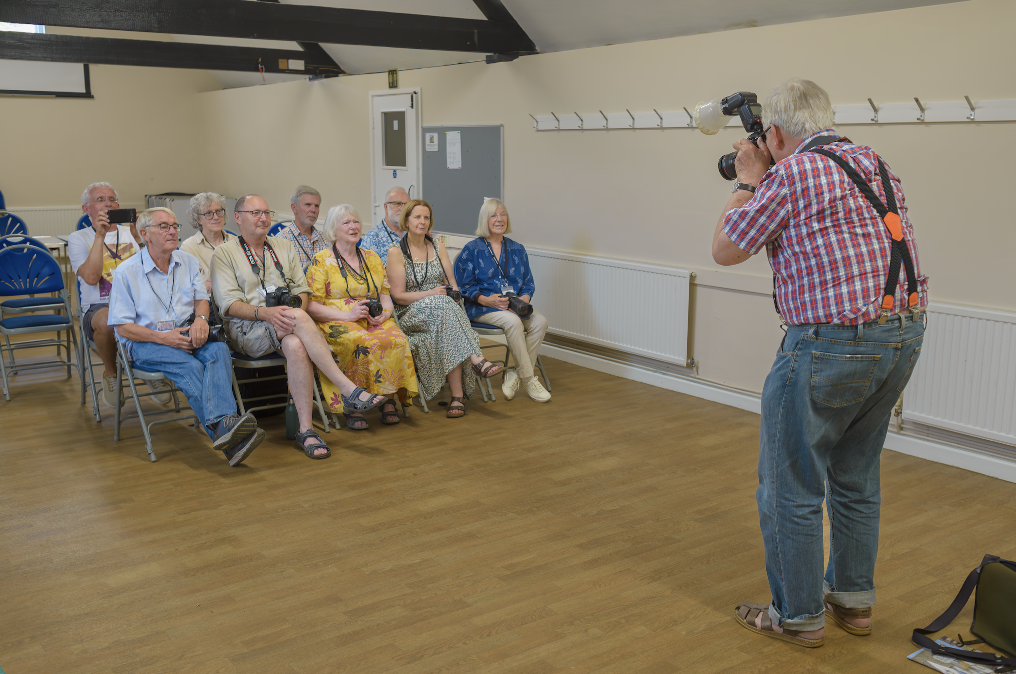 Martin Parr, camera and ring flash raised, photographs a row of seated photography enthusiasts holding their own cameras in a village hall.