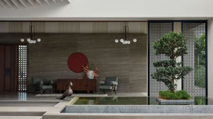A home in Tirupur, India with a beautiful porch featuring a koi pond, a bonsai tree, a pair of gray armchairs, a vase with bird of paradise, a red wall hanging, and a peacock statue