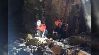 We see two researchers wearing hard hats with flashlights on them. They are in a dark cave. One is looking down at rocks and the other is looking off to the right.
