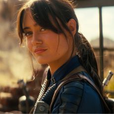 ella purnell smirks while wearing a blue suit and standing in the desert in fallout season 2