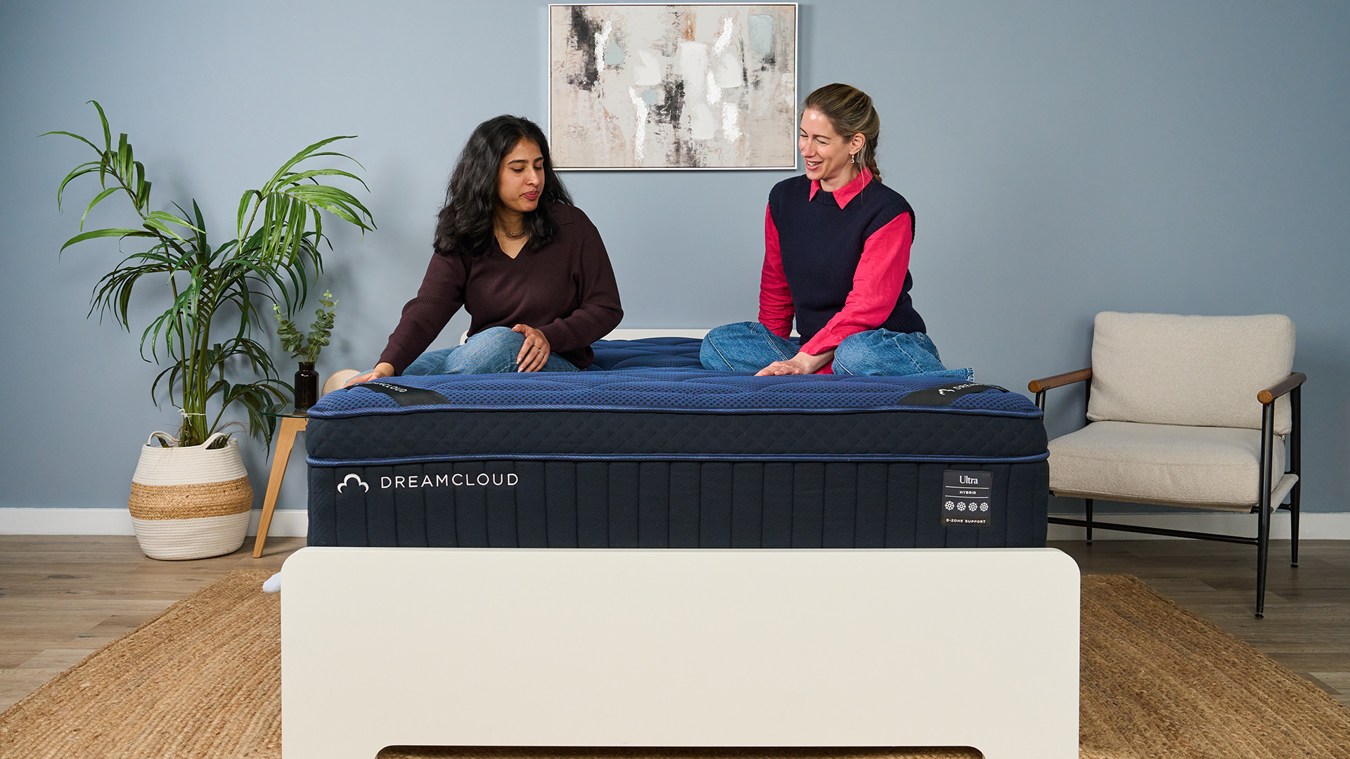 The image shows two mattress testers sitting on top of a navy blue mattress, placed on a white bed frame in a muted blue bedroom.