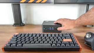 A person pressing the Copilot button on the front of the MSI Cubi NUC AI+ 2MG mini PC on a desk with a dual-monitor setup in the background and a mechanical keyboard and trackball mouse in the foreground