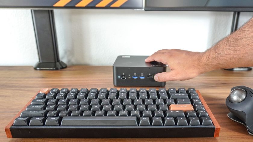 A person pressing the Copilot button on the front of the MSI Cubi NUC AI+ 2MG mini PC on a desk with a dual-monitor setup in the background and a mechanical keyboard and trackball mouse in the foreground
