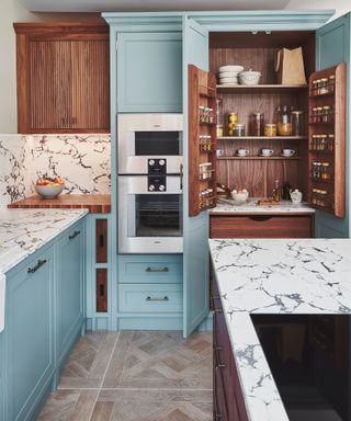 A kitchen with teal-painted cabinetry with integrated stainless steel ovens and walnut accents. The larder cupboard has walnut shelving, spice racks on the doors, and marble-lined surfaces. Marble countertops run throughout, complemented by a herringbone-patterned tile floor.