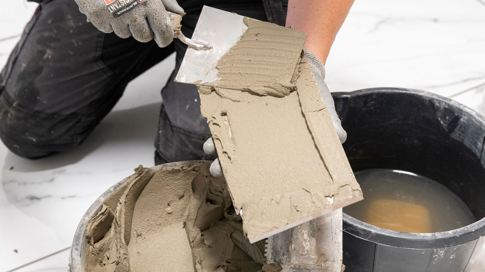 Man spreading tile adhesive with a bucket trowel onto the back of a tile trowel with a bucket of adhesive and bucket of water in foreground