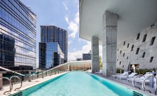 A pool area with a long pool, white loungers and shrubbery surrounded by glass buildings.