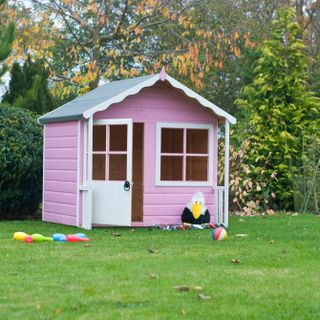 Kitty playhouse in a large garden, pink with grey roof, white door and trimmings, with toys scattered in front of it