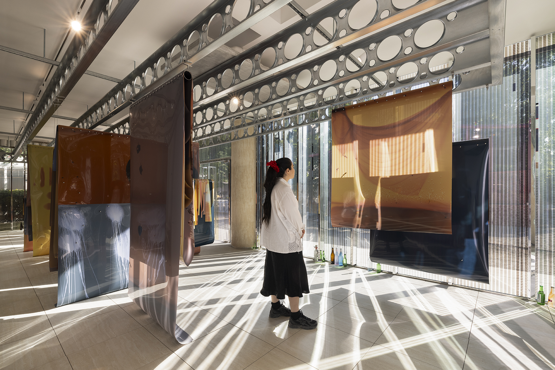 A woman studies large sheets of tinted photographic film hanging from steel beams in a sunlit gallery