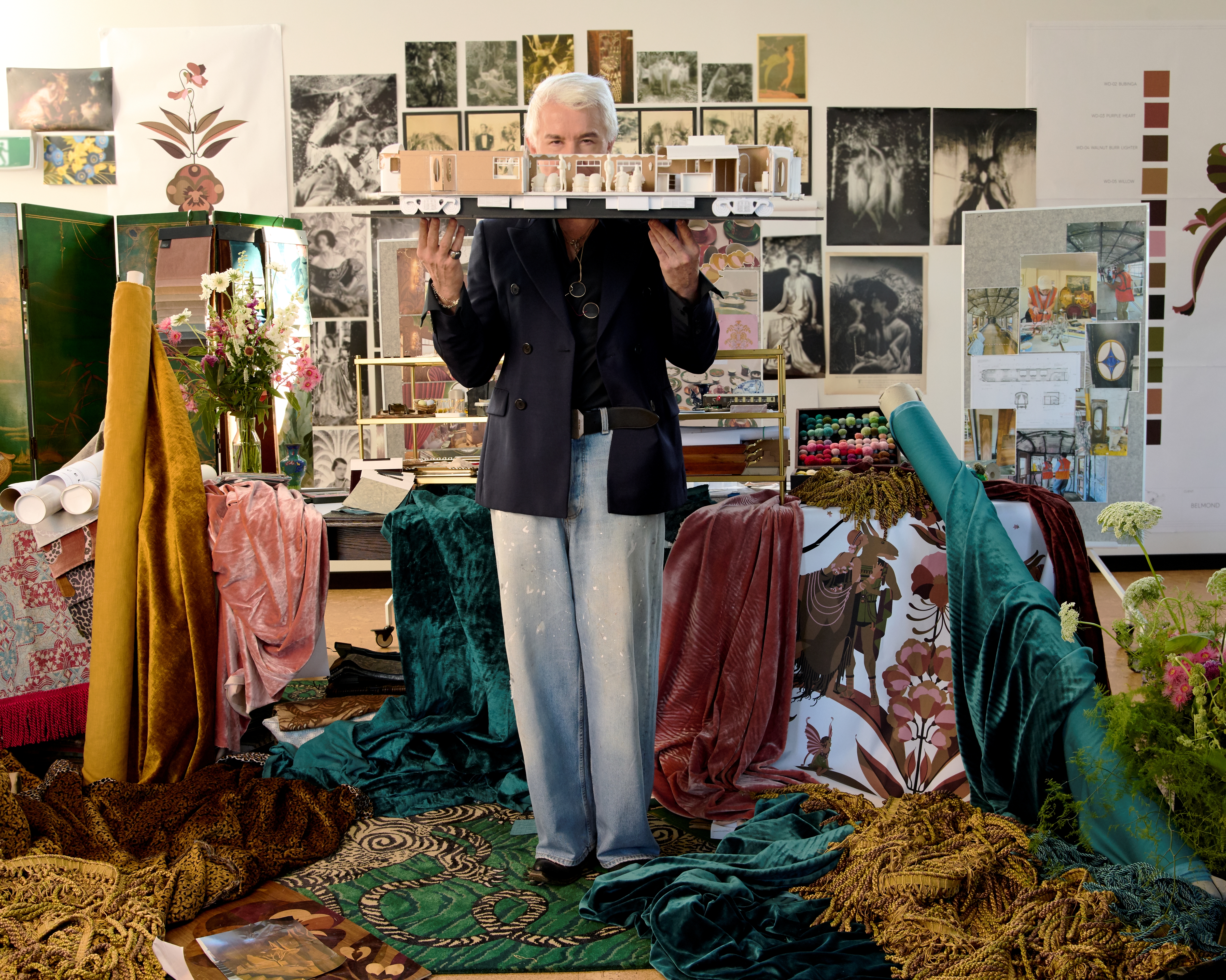 Baz Luhrmann in a textiles, posters, pictures, and sketches-filled studio stands with a model of a train in front of his face mid-air.