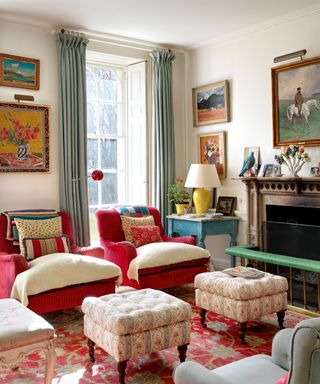 Living room in a country style with matching red upholstered armchairs and matching ottomans with patterned upholstery, large red and blue patterned rug, green curtains, colorful pictures and paintings on the wall, blue painted side table, yellow lamp shade