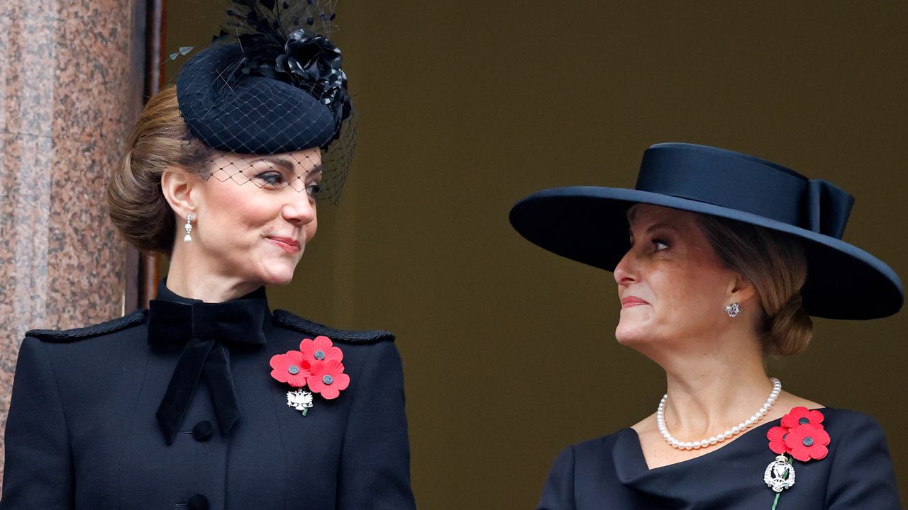 Kate Middleton wearing a black hat and coat turning to the right and smiling at Duchess Sophie, also wearing a black hat and coat with a red poppy brooch