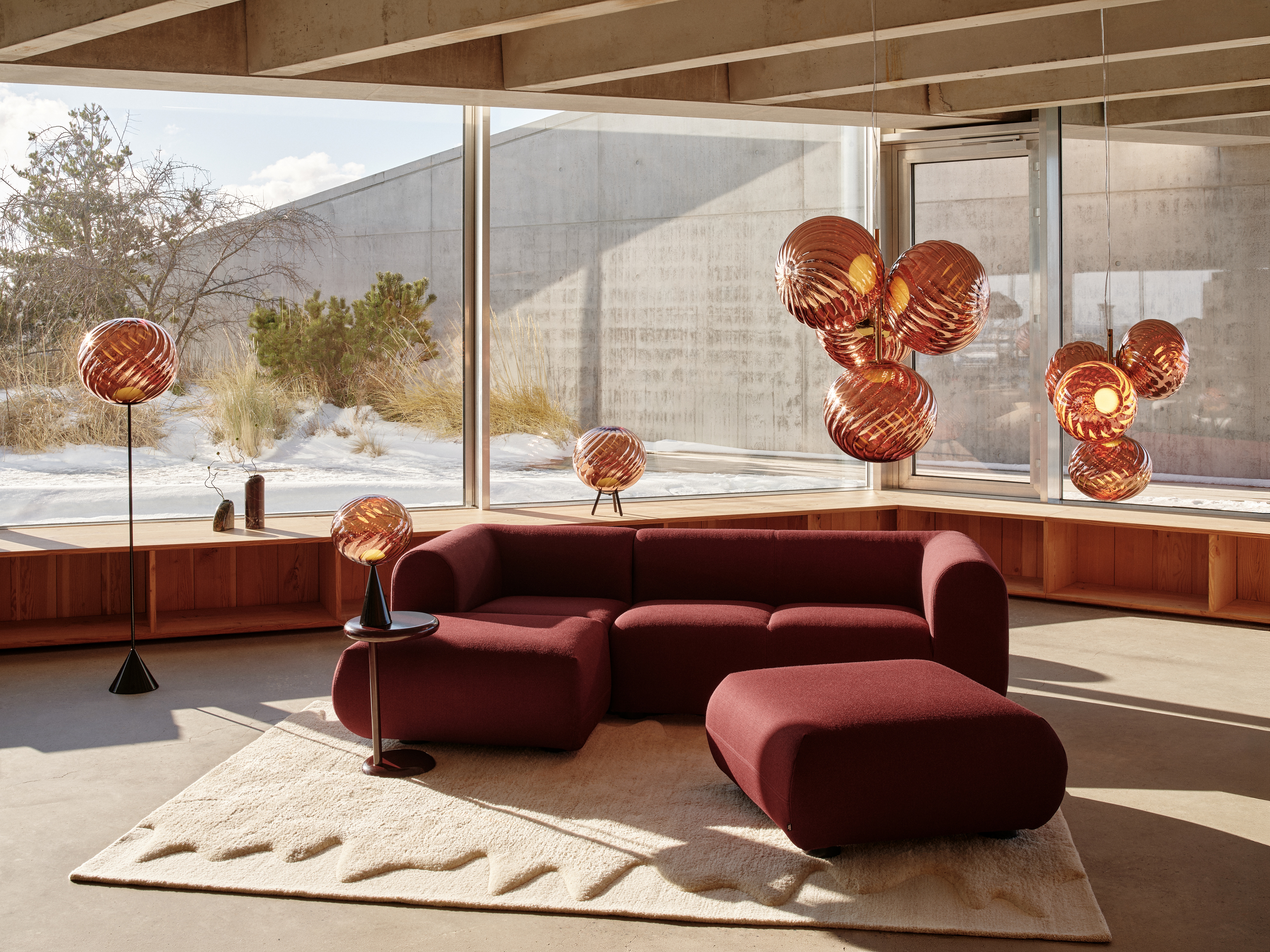 A sun-lit hotel lounge decorated with a chubby, cherry plum-red couch, pendant copper lighting, wooden ceilings, sandy floors, and floor-to-ceiling windows.