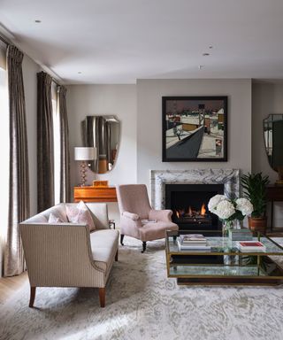 A stlish living room painted with a mid gray-beige, with mirrors and spotlights and a glass coffee table in front of a fireplace.
