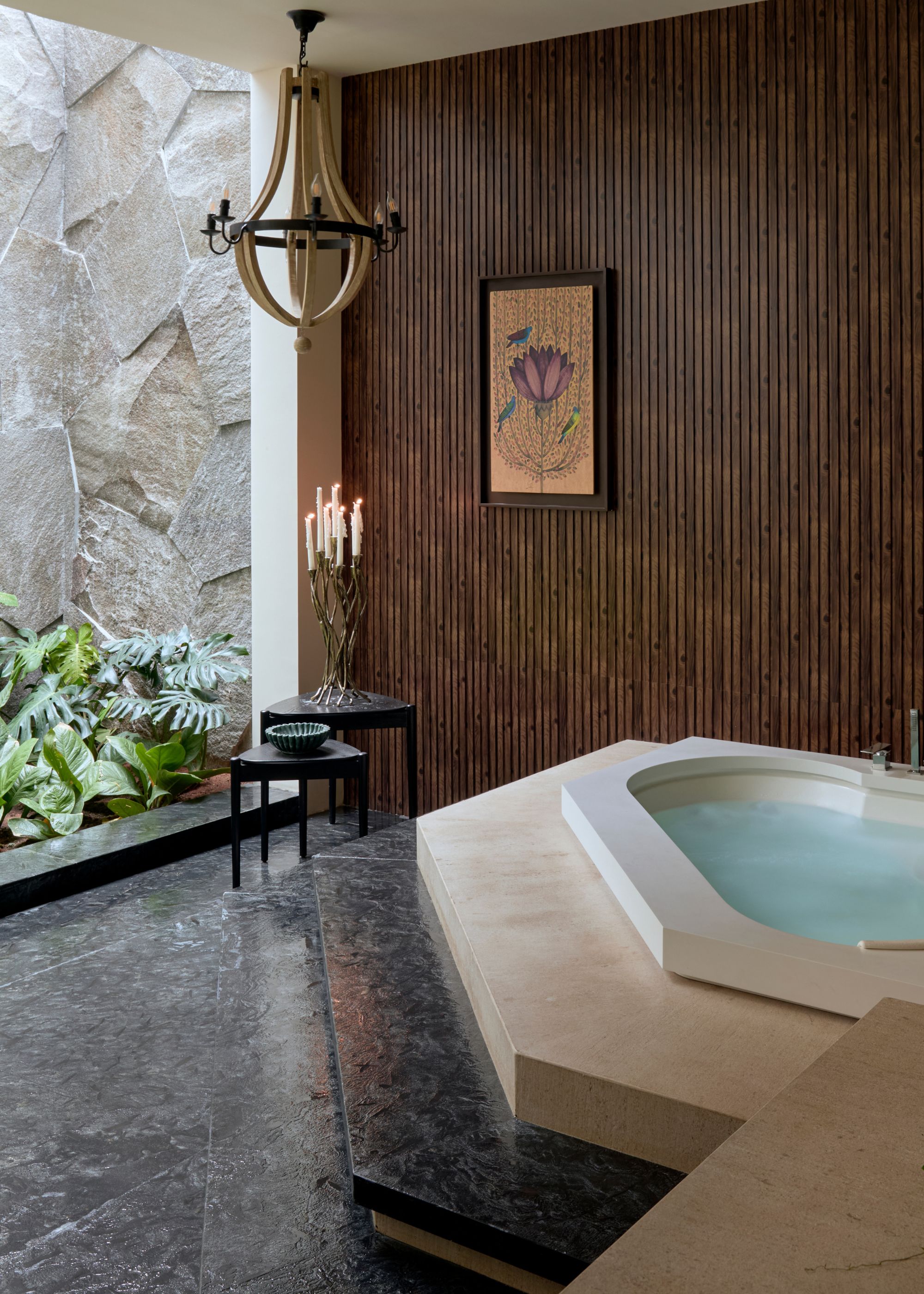 A spa bathroom with a tub, a candelabra with lit candles, a plant corner, a wood chandelier, and a framed print
