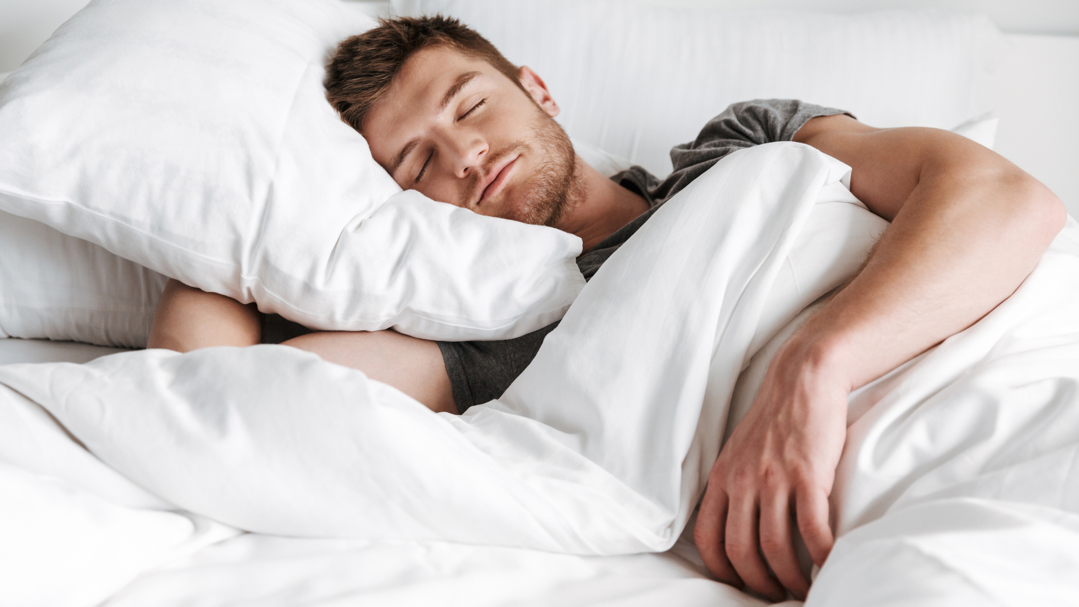 A man with short brown hair sleeps comfortably in a white bed