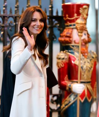 Kate Middleton wearing a white coat and waving in front of a huge nutcracker statue