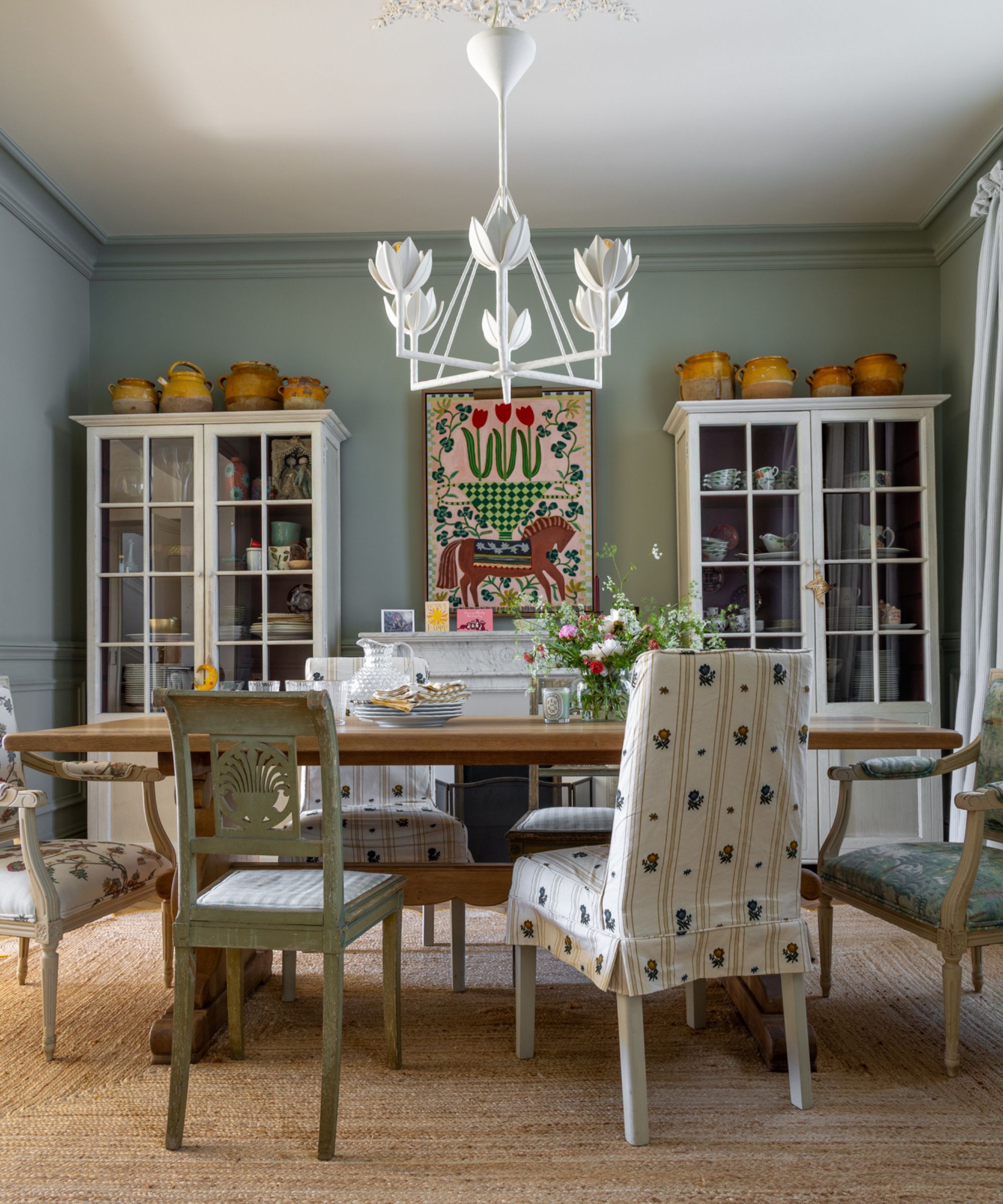 A dining room with sage green walls, a mix of upholstered dining chairs, a rustic wood table, twin glass-front cabinets, and a bold painting over the fireplace.
