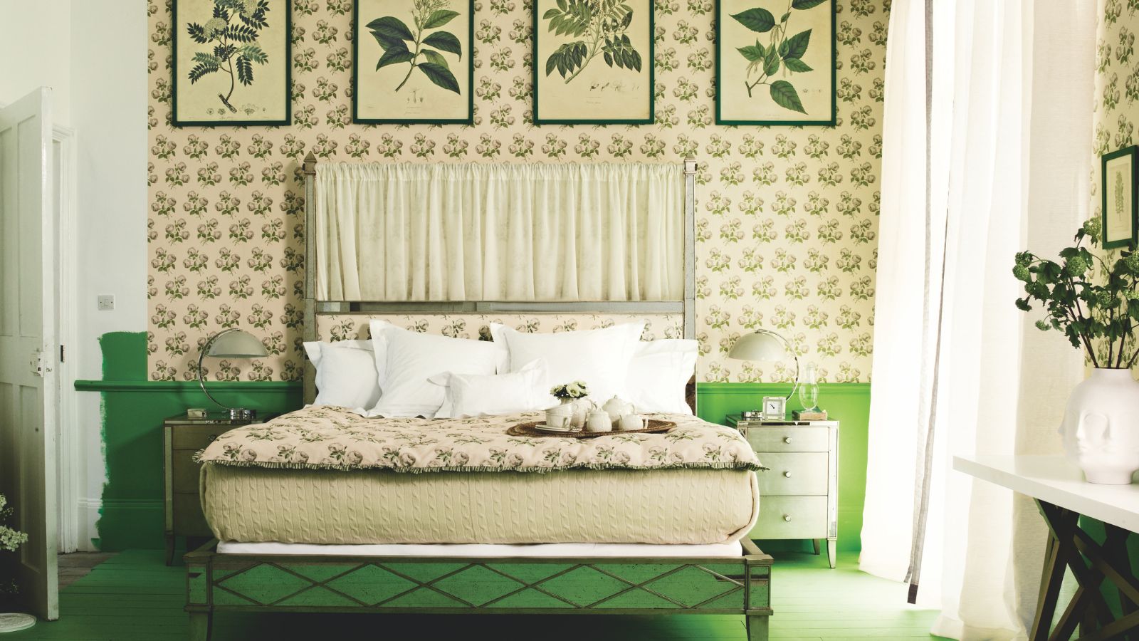 A green bedroom with floral wallpaper, botanical prints, a green bed with a patterned quilted blanket and white pillows, and a large window to the right with sheer white curtains. 