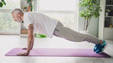 A man performs a plank exercise at home on an exercise mat. He is on his hands and toes, hips held high above the exercise mat, with his body held in a straight line from his shoulders to his heels. 