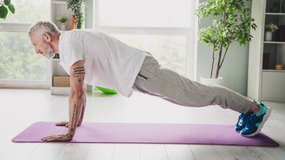 A man performs a plank exercise at home on an exercise mat. He is on his hands and toes, hips held high above the exercise mat, with his body held in a straight line from his shoulders to his heels. 