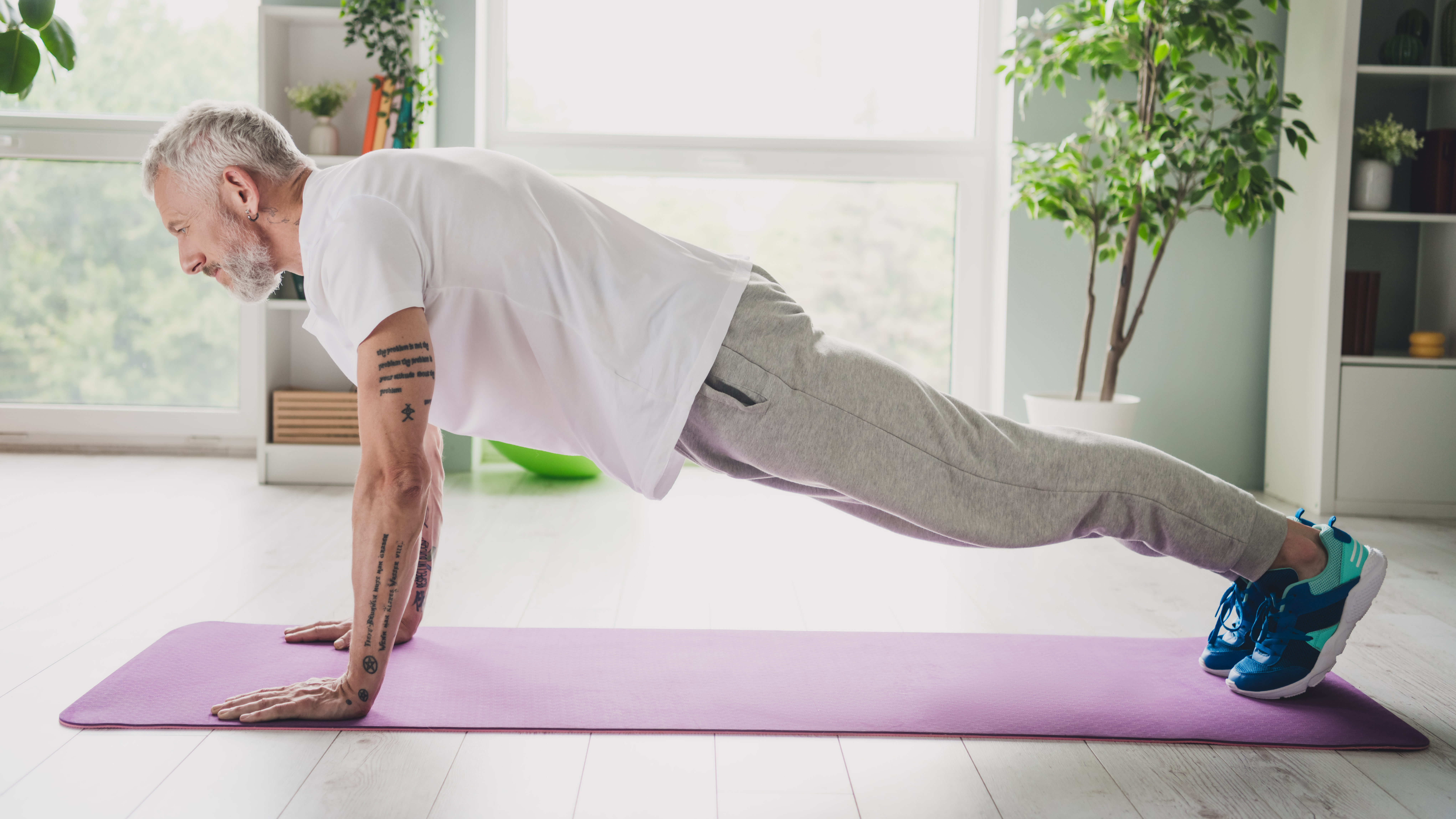 A man performs a plank exercise at home on an exercise mat. He is on his hands and toes, hips held high above the exercise mat, with his body held in a straight line from his shoulders to his heels. 