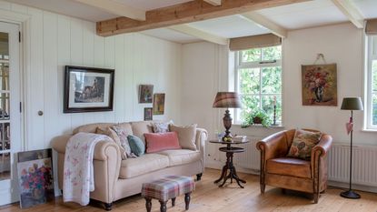 living room with white paneled walls cream sofa and leather tub chair bare floorboards and floral paintings