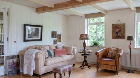 living room with white paneled walls cream sofa and leather tub chair bare floorboards and floral paintings