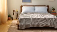 Striped brown and white Brooklinen Tencel sheets with blue pillows on a wooden double bedframe in a white bedroom with light wood flooring, jute rug, circular nightstand, books, and peach curtains.