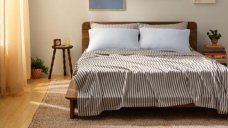 Striped brown and white Brooklinen Tencel sheets with blue pillows on a wooden double bedframe in a white bedroom with light wood flooring, jute rug, circular nightstand, books, and peach curtains.