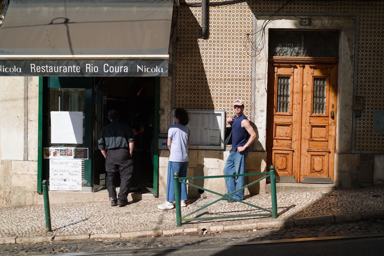 People standing outside a tiled Lisbon restaurant beside a wooden doorway in bright afternoon sunlight.