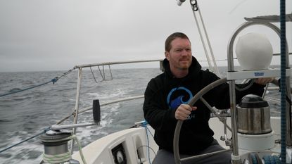 A man in a black hoodie stands behind the wheel of a boat