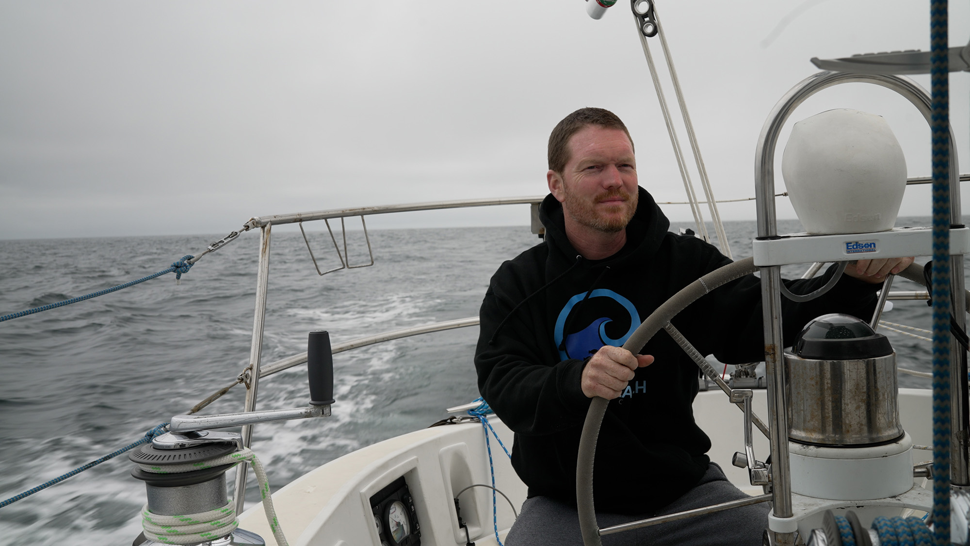 A man in a black hoodie stands behind the wheel of a boat