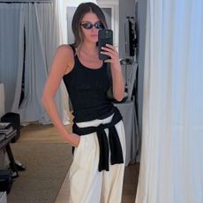 style influencer Coco Schiffer poses for a mirror selfie wearing black oval sunglasses, a black tank top, white relaxed pants, and a black sweater tied around her waist