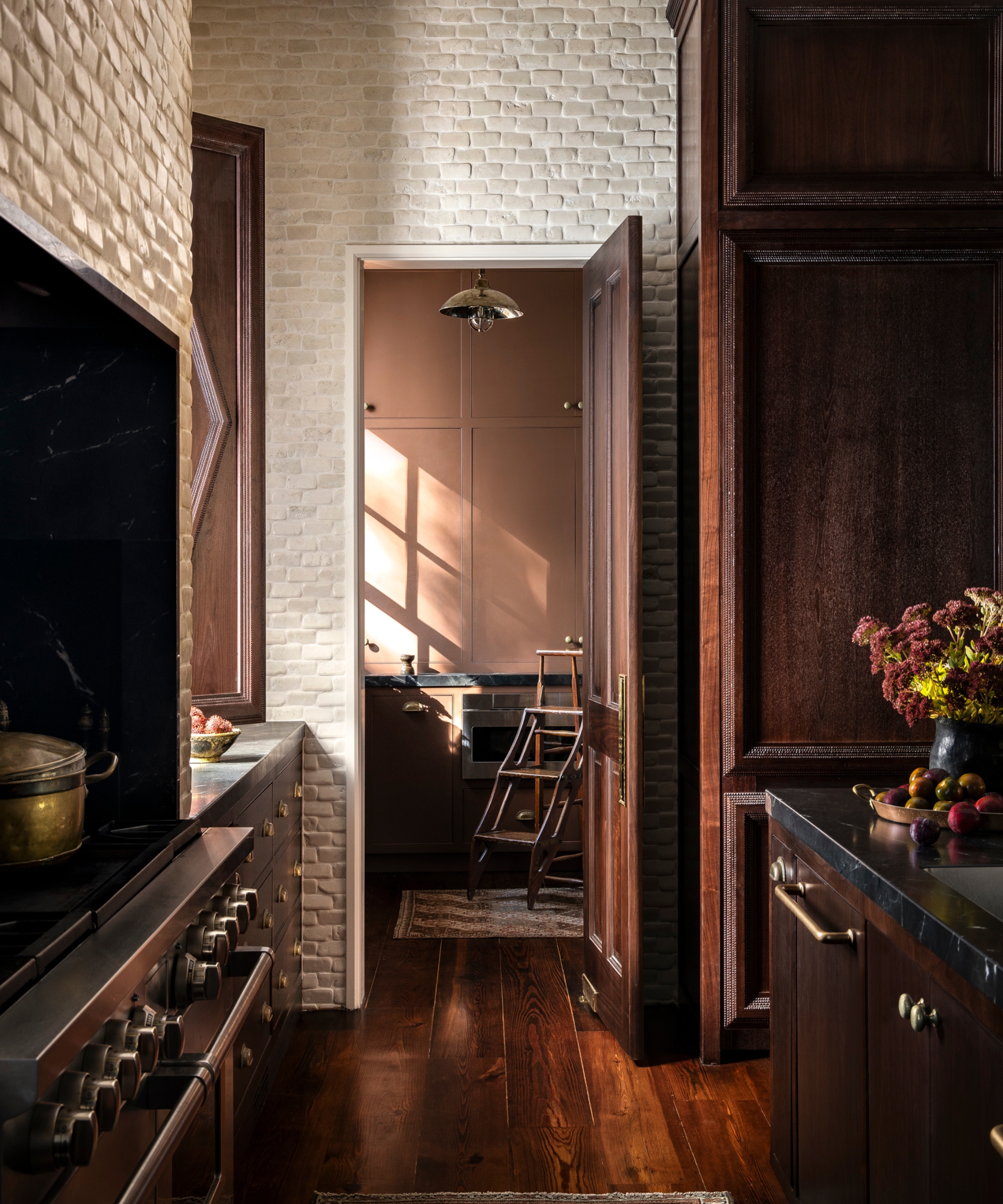 A moody kitchen with dark wood cabinets, white brick walls, and a door into a burnt orange-pink pantry