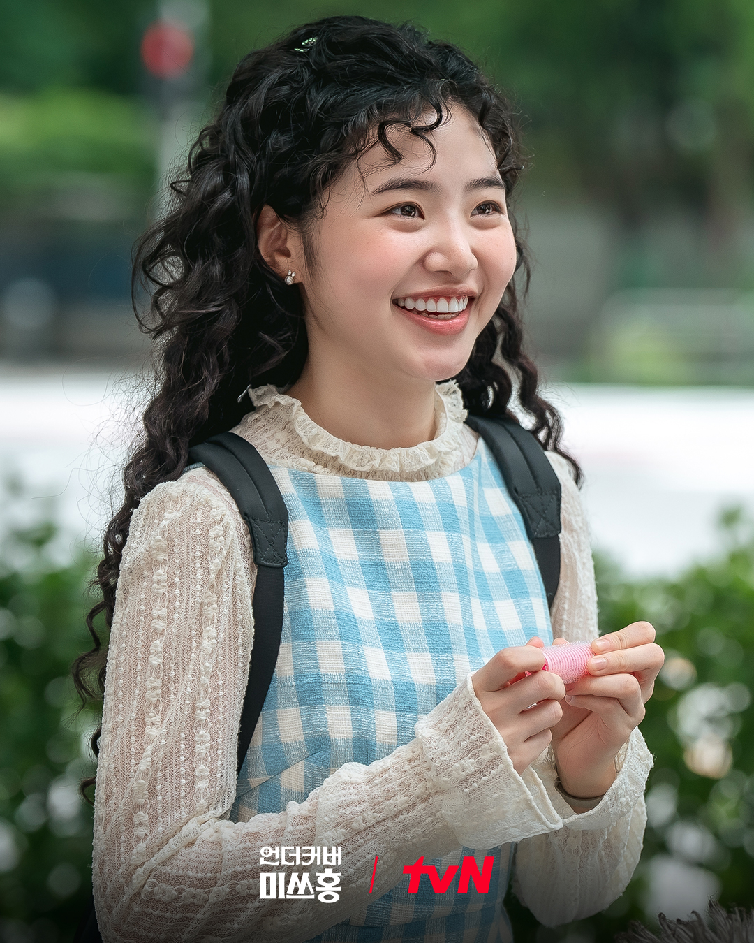 A woman in a blue checkered shirt, lace blouse, and backpack (Choi Ji-su as Kang Nara) smiles while standing in front of foliage, in the Netflix K-drama 'Undercover Miss Hong.'