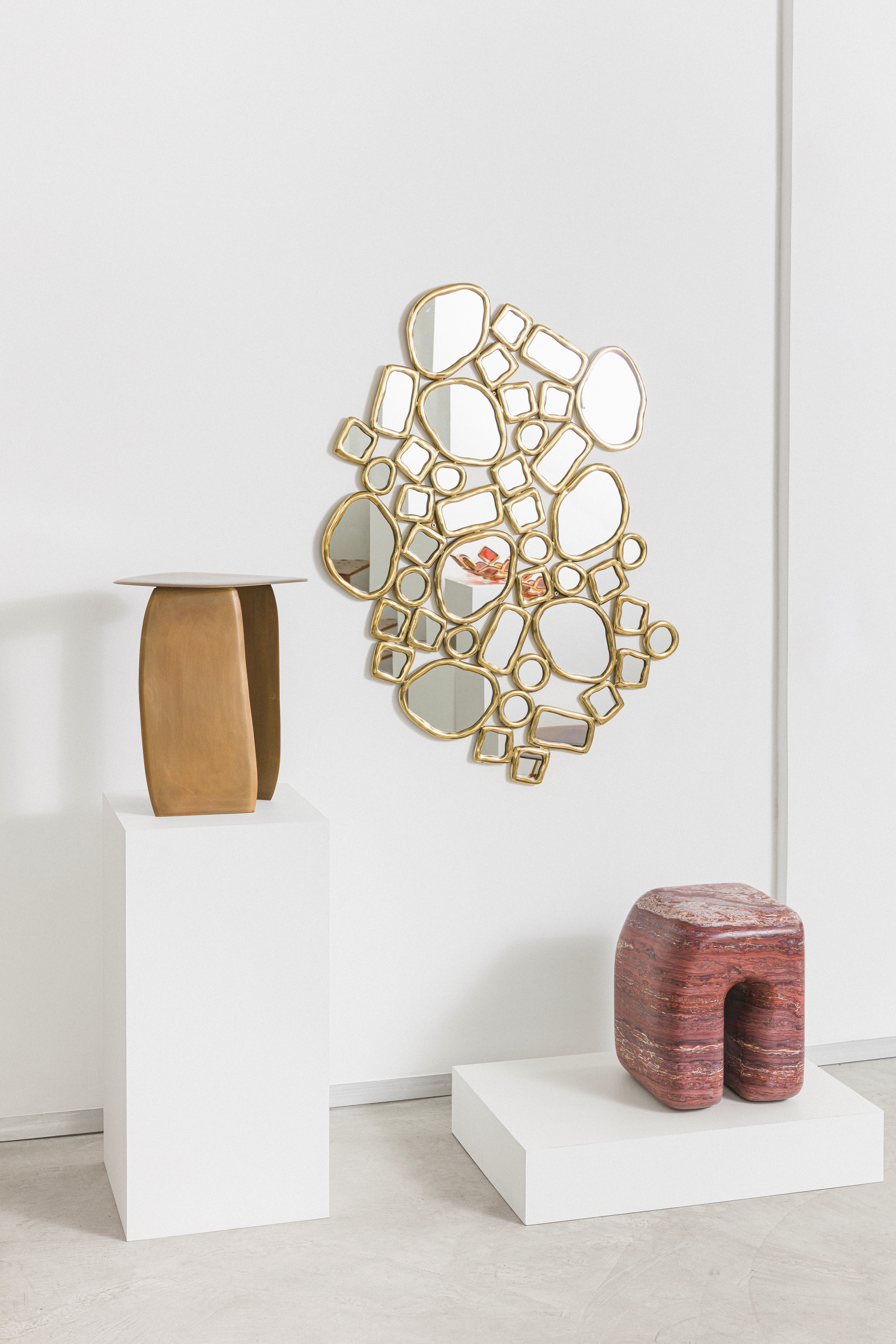 An installation view of a series of sculptural furniture pieces, including a red marble organic stool, a golden coffee table, and a multi-part mirror in brass.